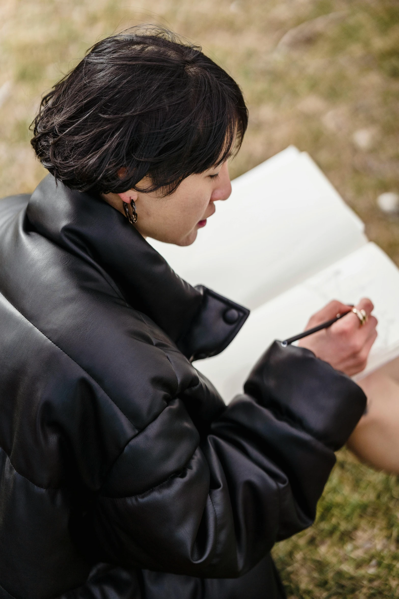 a woman sitting on the ground writing in a book, inspired by Ruth Jên, visual art, model wears a puffer jacket, short leather coat, field journal, close - up portrait shot