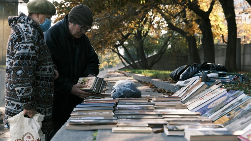 a couple of men standing next to a table full of books, by Attila Meszlenyi, unsplash, arte povera, city park, begging for alms, adi granov, fall season