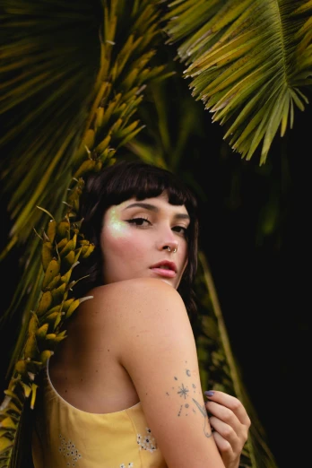 a woman in a yellow dress standing next to a palm tree, an album cover, inspired by Elsa Bleda, pexels contest winner, photorealism, with tattoos, on a planet of lush foliage, lights on, headshot profile picture