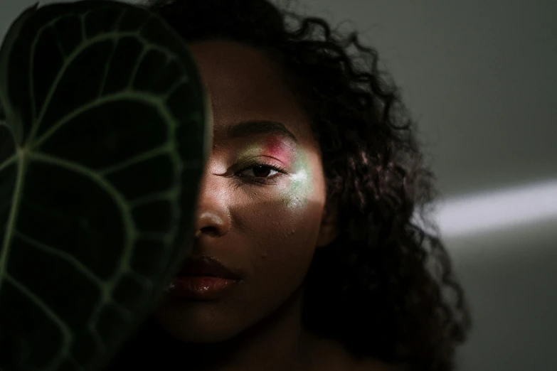 a woman holding a green leaf in front of her face, an album cover, by Adam Marczyński, trending on pexels, afrofuturism, light make up, her iridescent membranes, sza, looking into a mirror