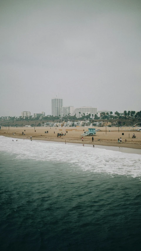 a group of people standing on top of a beach next to the ocean, a tilt shift photo, unsplash, !dream los angeles, big overcast, white buildings, medium format