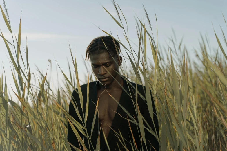 a man standing in a field of tall grass, an album cover, unsplash, brown skinned, non binary model, in a movie still cinematic, deep black skin