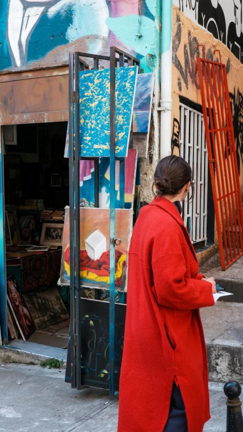 a woman in a red coat walking down a street, a gouache, pexels, process art, stands at a his easel, tel aviv street, spanish alleyway, side profile shot