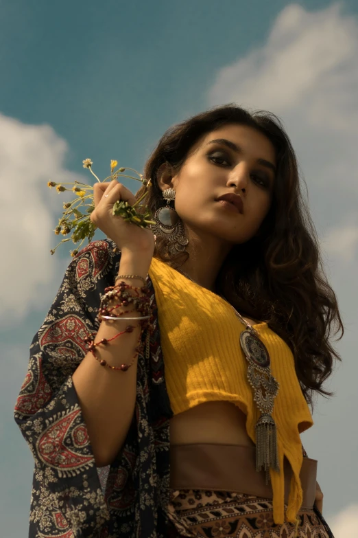 a woman in a yellow top holding a flower, by Riza Abbasi, trending on pexels, renaissance, tribal jewelry, at a fashion shoot, skies behind, square