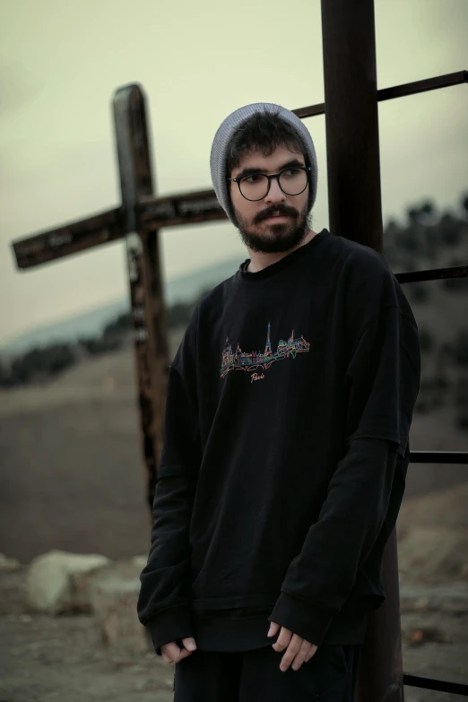 a man standing in front of a wooden cross, featured on reddit, photorealism, wearing sweatshirt, with glasses and goatee, hills in the background, gothic clothing