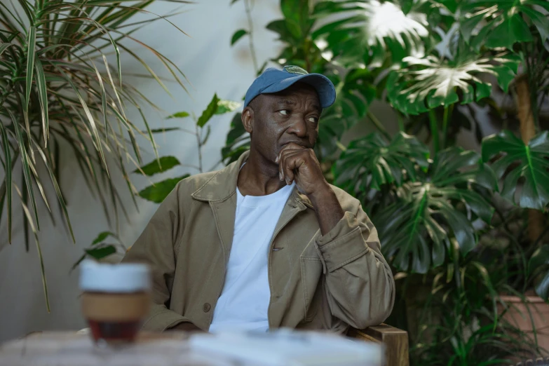 a man sitting at a table with a cup of coffee, amongst foliage, black man, wearing a baseball cap, ignant