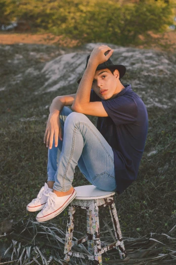 a man sitting on top of a stool in a field, by Adam Dario Keel, trending on pexels, androgynous face, young teen, jeans, canvas
