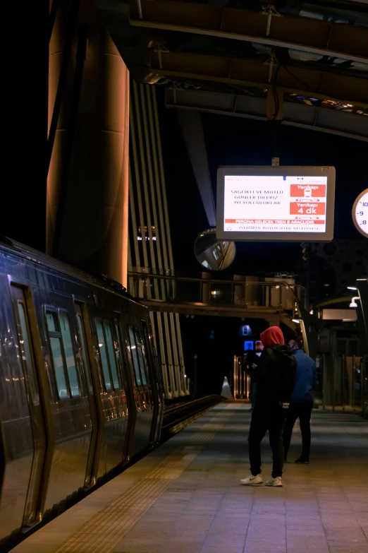 a couple of people standing next to a train, by Daarken, happening, huge viewscreen at front, nighttime!, orange line, electronic billboards