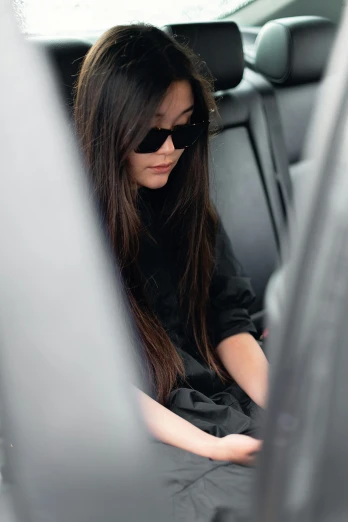 a woman sitting in the back seat of a car, black silky hair, dark shades, wearing black clothes, looking down