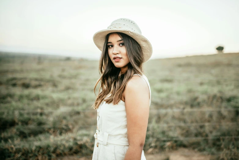 a woman standing in a field wearing a hat, by Arabella Rankin, pexels contest winner, hailee steinfeld, in front of white back drop, young asian woman, overalls