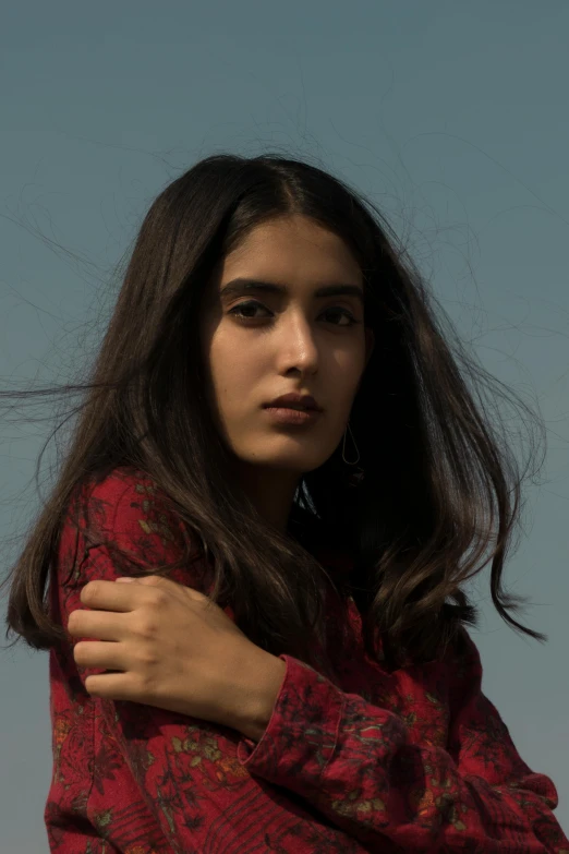 a woman standing in front of a blue sky, an album cover, by Maryam Hashemi, pexels contest winner, long hair and red shirt, still from a terence malik film, thick bushy straight eyebrows, 19-year-old girl