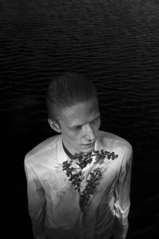 a man standing next to a body of water, a black and white photo, inspired by Gyula Basch, portrait of albino mystic, hedi slimane, lostus flowers, slicked back hair