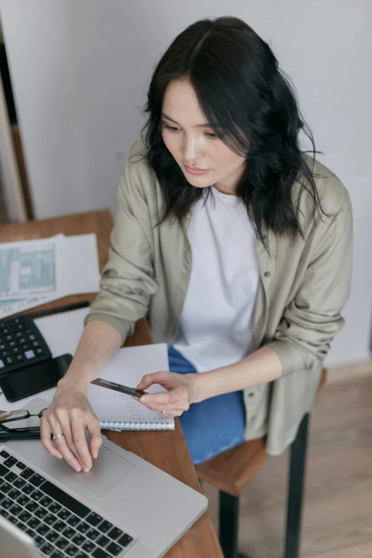 a woman sitting at a desk working on a laptop, by Nicolette Macnamara, wearing a linen shirt, holding pencil, asian woman, trending photo