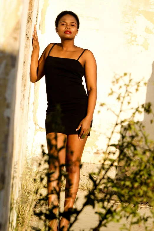 a woman in a black dress leaning against a wall, by Nadir Afonso, pexels contest winner, light skinned african young girl, full body:: sunny weather::, wearing a camisole and shorts, african domme mistress