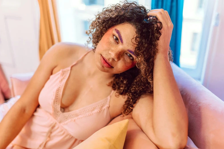 a woman sitting on top of a bed next to a window, a portrait, trending on pexels, renaissance, pastel pink skin tone, curls and curves, mixed race woman, multicoloured