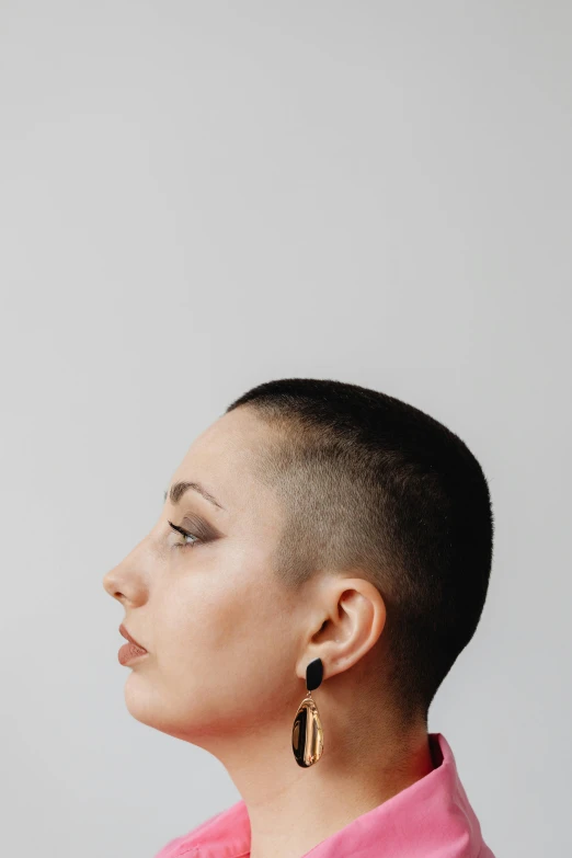 a woman with a shaved head wearing a pink shirt, trending on pexels, antipodeans, middle eastern skin, earring, profile pose, androgyny