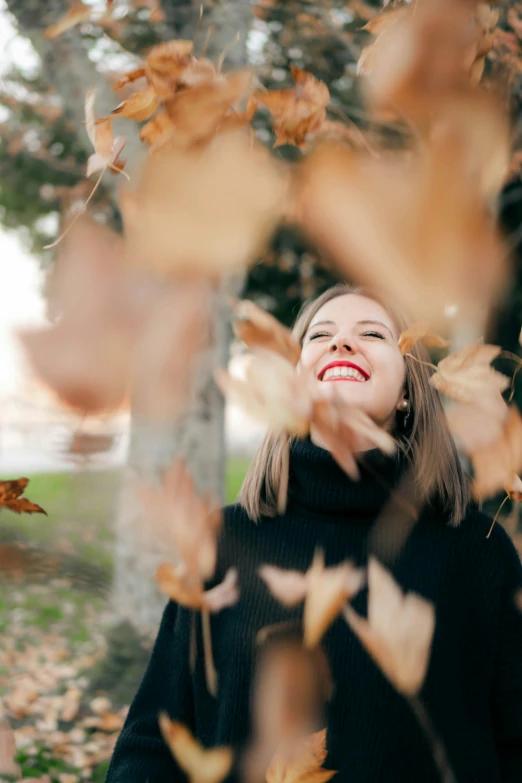 a woman standing next to a tree covered in leaves, pexels contest winner, smiling playfully, falling leaves, avatar image, 256435456k film