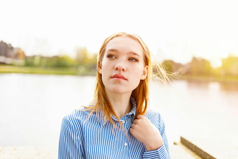 a woman standing next to a body of water, eleanor tomlinson, wearing stripe shirt, thoughtful expression, sunny sky