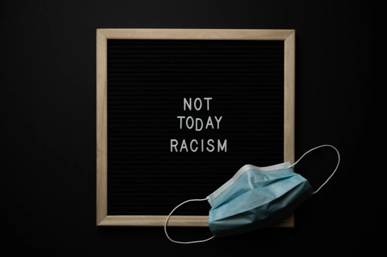 a mask on top of a sign that says not today racism, trending on unsplash, 1 6 x 1 6, abcdefghijklmnopqrstuvwxyz, compassionate, blackboard