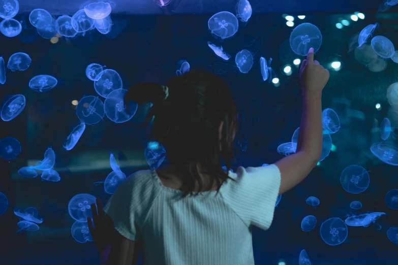 a woman standing in front of a display of jellyfish, inspired by Elsa Bleda, pexels contest winner, interactive art, tiny girl looking on, blue neon, kids, looking her shoulder