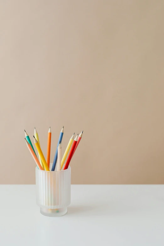 a glass filled with colored pencils on top of a table, by Nicolette Macnamara, on a pale background, minimalistic, small, promo image