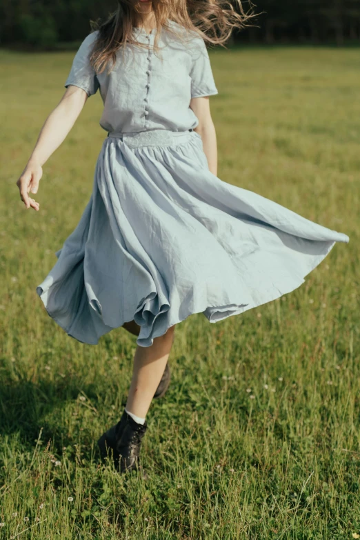 a woman walking across a lush green field, an album cover, by David Simpson, pexels, grey skirt, pale blue outfit, dancing gracefully, casual clothing