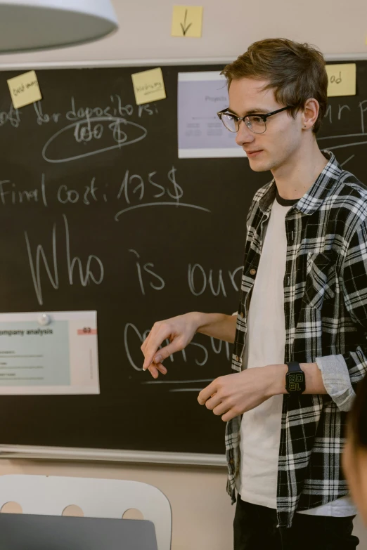 a man standing in front of a blackboard with writing on it, by Adam Marczyński, trending on unsplash, academic art, non binary model, standing in class, van allsburg, nerdy appearance