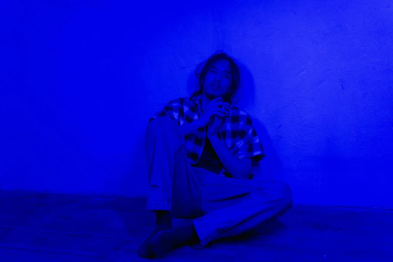 a woman sitting on the floor in front of a blue wall, an album cover, happening, blacklight aesthetic, frank dillane, low quality photo, on a checkered floor