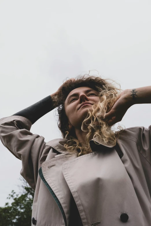 a woman standing on top of a lush green field, an album cover, trending on pexels, messy hair bedhead, woman is in a trenchcoat, on grey background, hands in her hair. side-view