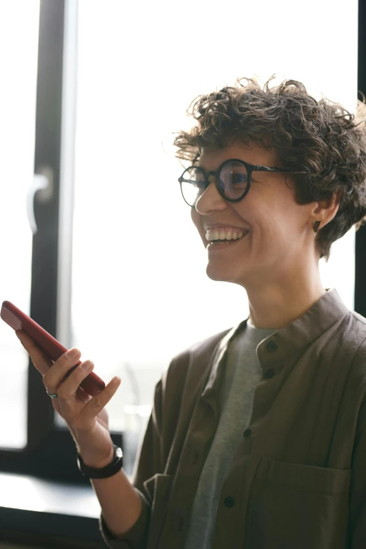 a woman standing in front of a window holding a cell phone, dark short curly hair smiling, wearing small round glasses, smiling at each other, customers