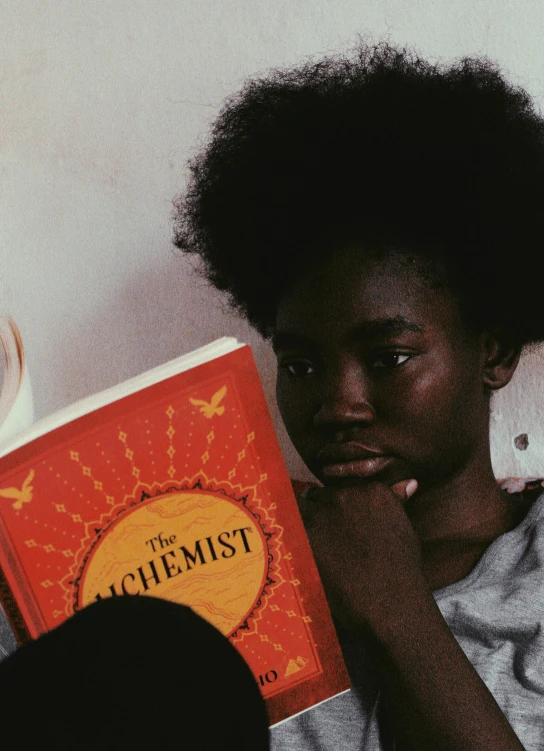 a person sitting on a bed reading a book, by Grace Clements, pexels contest winner, afrofuturism, human face with bright red yes, alchemist girl, with a white complexion, black teenage girl