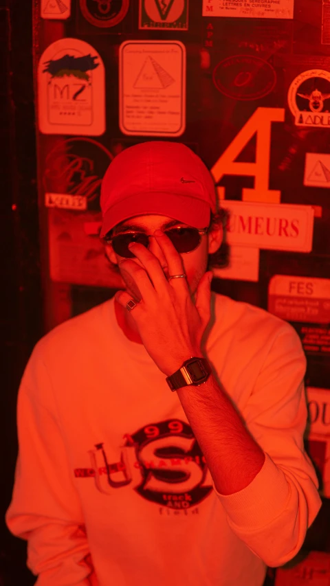 a man standing in front of a wall covered in stickers, an album cover, trending on pexels, les nabis, red cap with a capital m, dim red light, very smoky paris bar, he wears an eyepatch