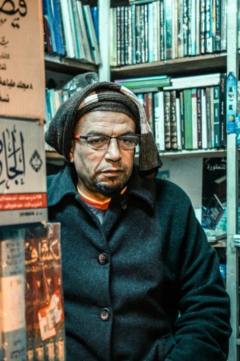 a man standing in front of a book shelf filled with books, an album cover, by Ahmed Yacoubi, pexels contest winner, les nabis, talaat harb square cairo, he‘s wearing a red scarf, an oldman, sitting down