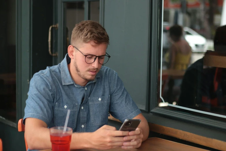 a man sitting at a table looking at his cell phone, by Carey Morris, pexels, with nerdy glasses and goatee, thin young male, gif, a blond