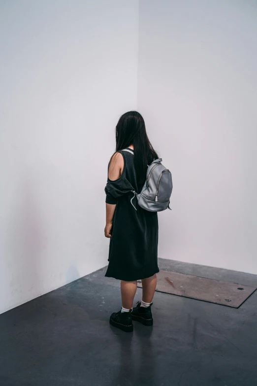 a woman standing in front of a white wall, inspired by Gao Cen, trending on unsplash, square backpack, metallic neoprene woman, exhibition, wearing a grey robe