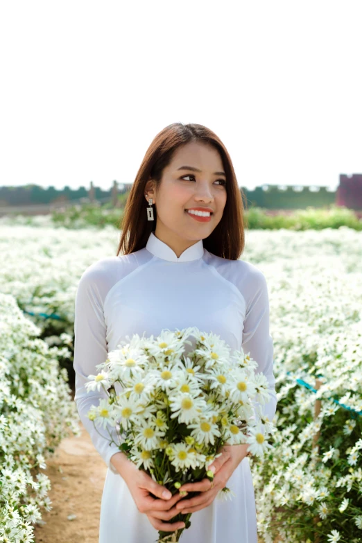 a woman in a white dress holding a bunch of flowers, inspired by Ruth Jên, pexels contest winner, ao dai, chamomile, while smiling for a photograph, long sleeves
