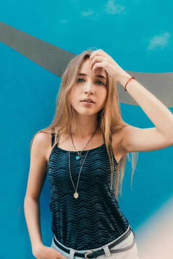 a woman standing in front of a blue wall, an album cover, inspired by Julia Pishtar, trending on pexels, graffiti, she is wearing a black tank top, jewelry, very grainy image, young with long hair