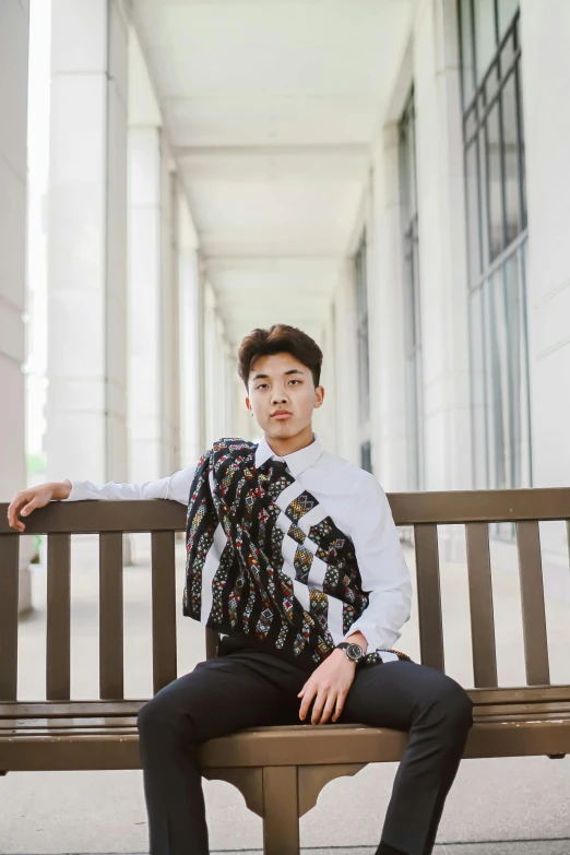 a man sitting on top of a wooden bench, inspired by Fei Danxu, unsplash, realism, patterned clothing, graduation photo, beautiful androgynous prince, in front of white back drop