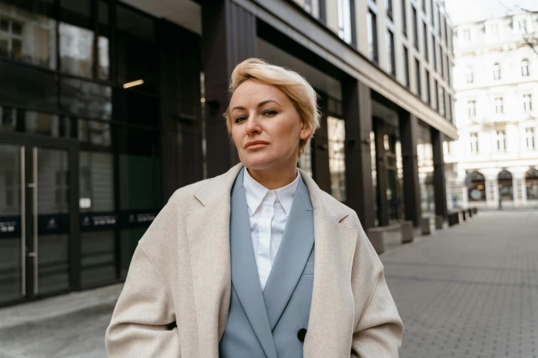 a woman standing on a sidewalk in front of a building, trending on pexels, trench coat and suit, blonde swedish woman, thumbnail, ukrainian