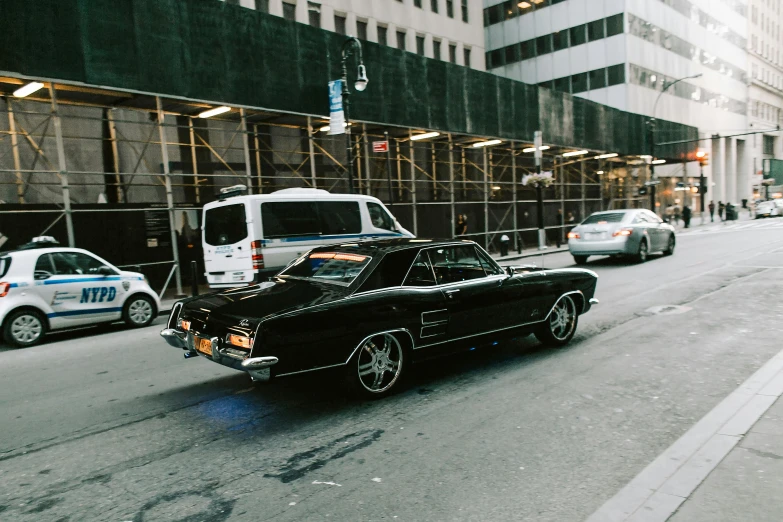 a black car driving down a street next to tall buildings, inspired by Elsa Bleda, pexels contest winner, standing in front of lowrider, new york streets, the last v 8 interceptor, 🚿🗝📝