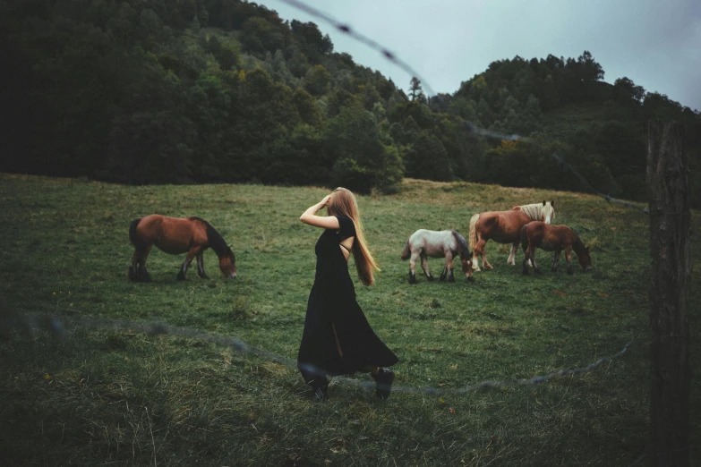 a woman in a black dress standing in a field with horses, pexels contest winner, julia hetta, feral languid woman, various posed, in the yard