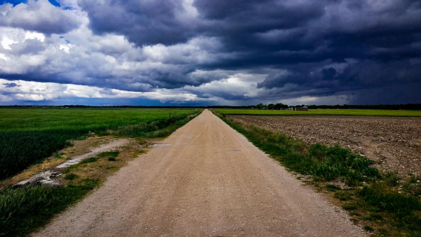 a dirt road in the middle of a field under a cloudy sky, by Daniel Seghers, unsplash, realism, with dark clouds in the sky, traveling in france, wide greenways, outside a storm rages