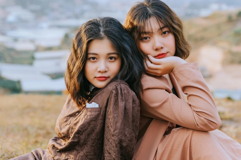 two women sitting next to each other in a field, trending on pexels, realism, young adorable korean face, brown clothes, malaysian, ad image