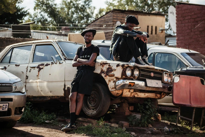 two men standing on top of an old car, by Lee Loughridge, pexels contest winner, afrofuturism, wearing black old dress and hat, standing in township street, sitting down, promotional image