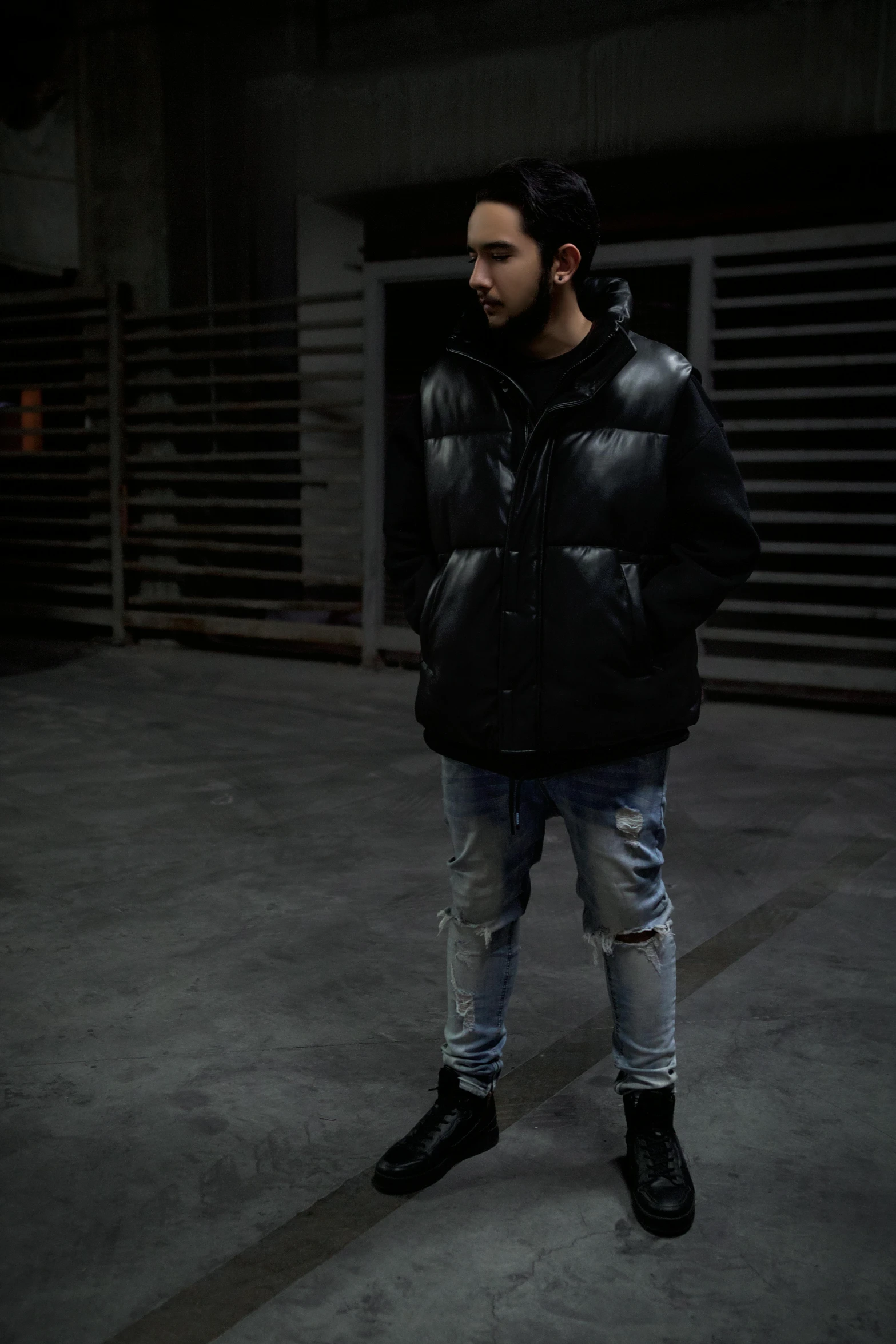 a man in a black jacket standing in a garage, an album cover, inspired by Amir Zand, unsplash, wearing jeans and a black hoodie, reflective puffy coat, dark. no text, model is wearing techtical vest