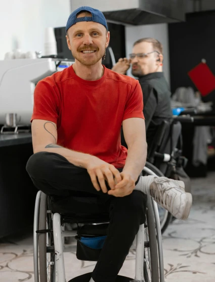 a man in a red shirt is sitting in a wheelchair, a colorized photo, trending on pexels, proud smirk, 😭🤮 💔, in a workshop, wearing pants and a t-shirt