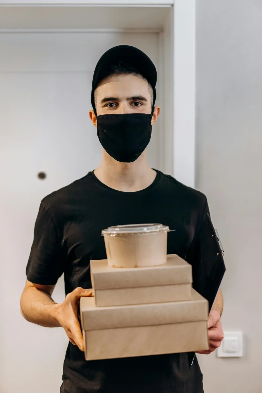 a man wearing a face mask holding a stack of boxes, wearing a black shirt, smol, balaclava, caucasian