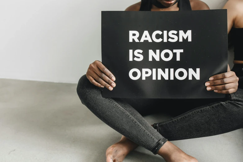 a woman sitting on the floor holding a sign that says racism is not opinion, a black and white photo, trending on unsplash, rayonism, background image, wearing a brown, a person standing in front of a, featured art