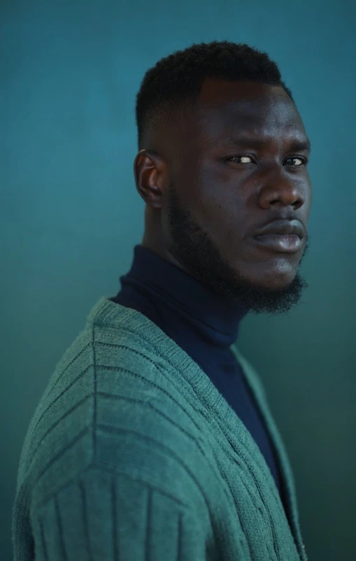 a man standing in front of a blue background, an album cover, inspired by David Bailly, trending on pexels, grey skin, wearing a green sweater, portrait. 8 k high definition, moody light