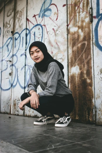a woman squatting in front of a graffiti covered wall, inspired by Naza, pexels contest winner, grey sweater, ((portrait)), muslim, portrait androgynous girl
